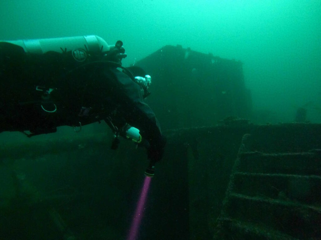 Bild eines Tauchers mit Lampe im trüben Wasser über einem Schiffswrack.